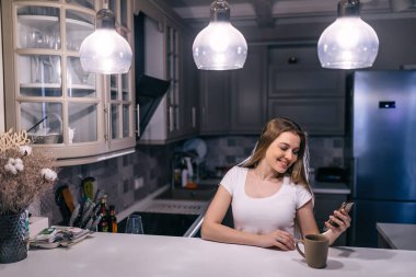 Gülümseyen genç bir kadın akşamları evdeki mutfak masasında oturmuş video görüşmesini kullanıyordu. Kahve içmek.