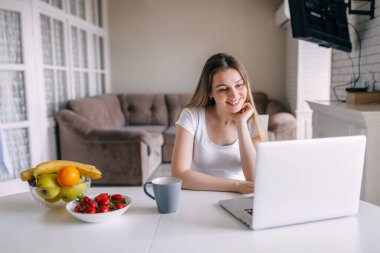 Bir fincan kahveyle gülümseyen genç bir kadın evdeki mutfakta dizüstü bilgisayarla video sohbeti yapıyor.