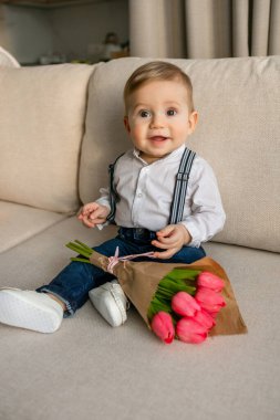 Beyaz tişörtlü, pantolon askısı takmış, pembe çiçekli koltukta oturan tatlı bir erkek bebek..