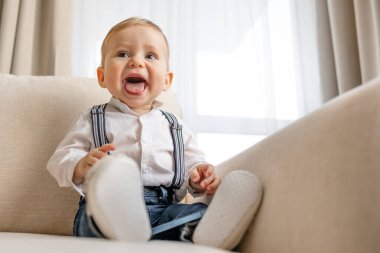 Evdeki bej bir koltukta oturan neşeli bir bebek, beyaz bir gömlek ve pantolon askısı giymiş, parlak bir odada mutlu bir şekilde gülümsüyor..