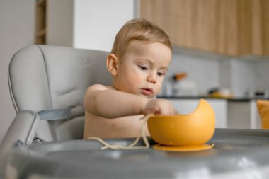 Yüksek bir sandalyede oturan sevimli bebek modern bir mutfakta sarı bir kaseden yemek yiyor. Bebek evde yemek yiyor..