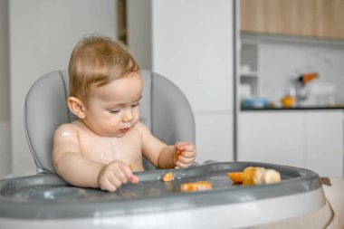 Güzel bebek yüksek sandalyede yemek yiyor, rahat bir evde meyve dilimleri yiyor..