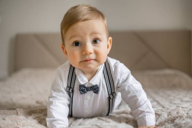 Papyon takan ve pantolon askısı takan sevimli bir erkek bebek yatakta emeklerken görülüyor, merak ve masumiyet gösteriyor..