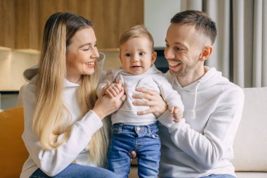 Gülümseyen ebeveynler evdeki kanepede otururken bebeklerini tutuyorlar. Aile içeride birlikte vakit geçirmekten hoşlanıyor..