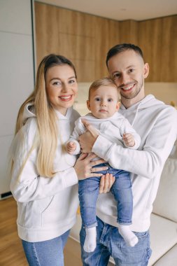 Beyaz kapüşonlu ve mavi kotlu rahat bir evde bebeğini kucağında tutan gülümseyen ebeveynler. Mutlu aile anı kapalı alanda yakalanır..