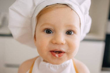 Aşçı gibi giyinmiş, yüzünde un olan, mutfakta ışıl ışıl gülümseyen bir bebeğin yakın çekimi..