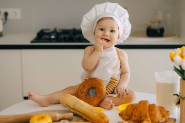 Aşçı gibi giyinmiş sevimli bir bebek mutfak masasında oturuyor, taze pişmiş kruvasan ve ekmeğin tadını çıkarıyor. Sevimli ve iç açıcı sahne aile ve yemek temaları için mükemmel..