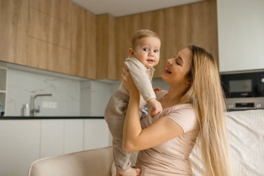Neşeli anne bebeğini modern bir mutfak ortamında kucağına alıp kaldırıyor. Anne ve çocuk bağı..