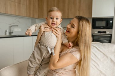 Neşeli bir anne, mutlu bebeğini modern, rahat bir mutfakta tutuyor. Anne bebeği tutarken gülümsüyor, sıcak ve sevgi dolu bir atmosfer yaratıyor..