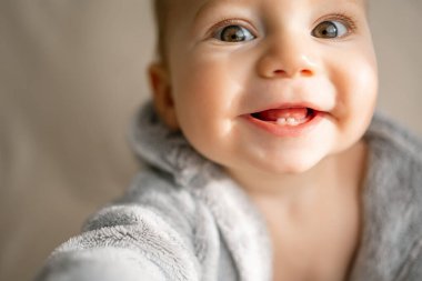 Gülümseyen bir bebeğin sevimli yakın çekimi dişlek bir sırıtışı gösteriyor ve yumuşak gri bir bornoz giyiyor..