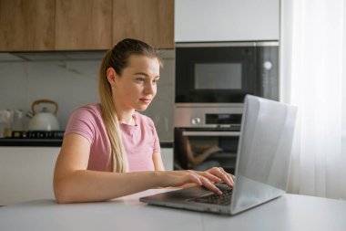 Modern bir mutfakta dizüstü bilgisayarıyla çalışan genç bir kadın. Uzaktan kumandalı çalışmalarına ve esnekliğine odaklanmış durumda. Dekor, doğal ışığı olan çağdaş bir mutfak..