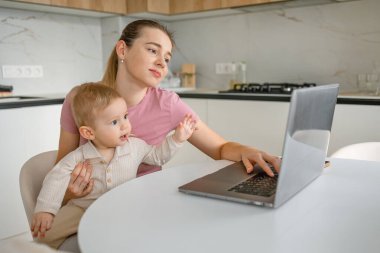 Anne kucağında bebekle evde birden fazla iş yapıyor, dizüstü bilgisayar kullanıyor, iş hayatının dengesini gösteriyor..