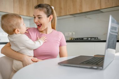 Modern bir mutfak ortamında dizüstü bilgisayarla uğraşırken bebeğini tutan mutlu çalışan bir anne..