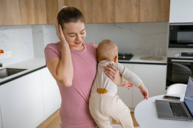 Yorgun ve genç bir anne bebeğini bebek bezi lekesiyle evden çalışırken modern bir mutfakta tutuyor..