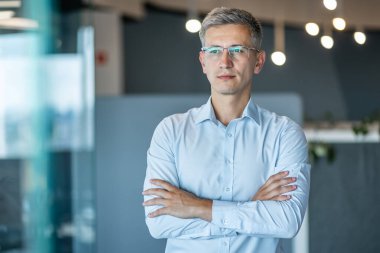 Modern bir ofiste kollarını kavuşturmuş kendine güvenen bir iş adamının portresi. Gözlüklü ve açık mavi gömlekli bir işadamı liderlik ve kararlılık hissi yayıyor..