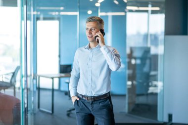 Açık mavi tişört giyen kendine güvenen bir işadamı modern ofis ortamında cam duvarlı bir odada akıllı telefonuyla konuşur. iletişim, başarı ve modern iş konsepti.