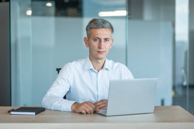 Beyaz gömlekli, profesyonel, orta yaşlı bir işadamı modern ofis ortamında bir masada oturuyor ve özgüvenle dizüstü bilgisayarı üzerinde çalışıyor..