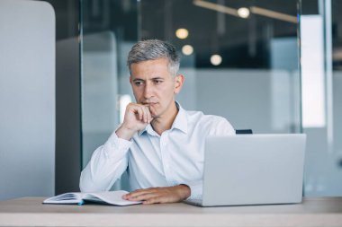 Düşünceli bir işadamı, modern bir ofiste, bir dizüstü bilgisayar ve defterle çevrili bir masada oturuyor ve stratejiyi yansıtıyor.