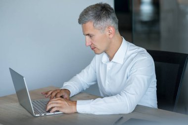 Beyaz gömlekli olgun bir iş adamı modern ofisindeki masasında dizüstü bilgisayara konsantre olur. şirket işleri, teknoloji ve iş görevleri.
