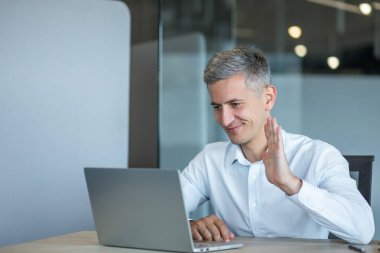 Modern bir ofis ortamından gelen bir video konferansında, profesyonel bir adam bilgisayarına el sallıyor ve çevrimiçi bir toplantı sırasında arkadaşça bir etkileşim gösteriyor..