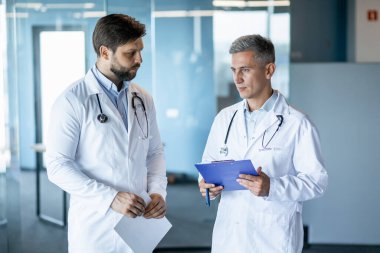Beyaz laboratuvar önlüklü iki doktor modern ve parlak bir hastane ortamında tıbbi kayıtları tartışıyorlar. Sağlık uzmanları arasında tıbbi uzmanlık ve iletişim.