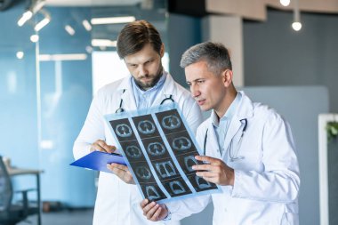 Beyaz önlüklü iki profesyonel doktor modern bir hastane ofisinde röntgen taramalarını dikkatle inceliyor. Tıbbi görüntülerle ilgili bulgular ve içgörüler tartışılıyor.