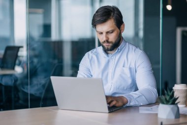 Açık mavi gömlekli sakallı bir adam modern ofis ortamında dizüstü bilgisayarda yoğun bir şekilde çalışır. Konsantrasyon ve verimlilik kavramı..
