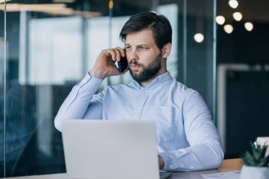 Mavi tişörtlü odaklanmış bir işadamı, modern bir ofis alanında dizüstü bilgisayarı üzerinde çalışırken telefonla konuşuyor..