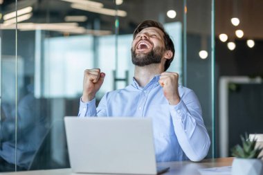 Şen şakrak bir işadamı ofis masasında otururken yumruklarını kaldırıyor. başarı, başarı ve pozitif enerji