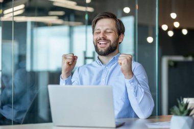 Mavi gömlekli sakallı bir adam modern ofis ortamında dizüstü bilgisayarın başında oturarak başarıyı kutluyor. neşe ve başarı, iş başarısını yansıtıyor.
