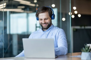 Modern bir ofiste dizüstü bilgisayarıyla çalışırken kulaklık takan neşeli bir adam gülümsüyor. üretkenlik, işyeri mutluluğu ve modern teknoloji.