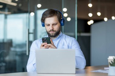 Kendine güvenen iş adamı kulaklık takıyor, modern ofis ortamında akıllı telefon ve dizüstü bilgisayar kullanıyor. Teknolojiye ve iş verimliliğine odaklanmış. iş, teknoloji ve konsantrasyon.