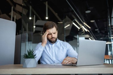 Masasında oturan yorgun bir işadamı eliyle başını tutuyor, acı ve hüsranı hissediyor. Modern ofis ortamı, bir dizüstü bilgisayar kullanarak.