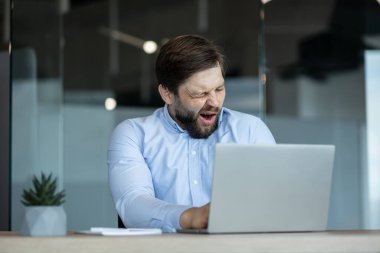 Mavi gömlekli sakallı bir adam bir masada oturuyor, dizüstü bilgisayarının önünde esniyor. Yorgunluk ve uzun çalışma saatlerinde odaklanmanın zorlukları.
