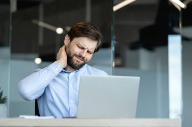 Ofisindeki masasında oturan bir iş adamı dizüstü bilgisayarı üzerinde çalışırken boynu ağrıyor. Uzatılmış bilgisayar kullanımı ve profesyonel ortamda kötü duruş ile ilgili rahatsızlıklar.