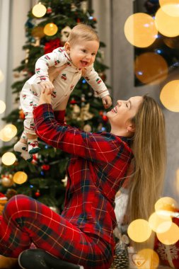 Neşeli bir anne, neşeli bebeğini güzel dekore edilmiş bir Noel ağacının yanında kaldırarak neşeli ruhu yakalıyor. Sıcak ışıklar ve rahat pijamalar tatil kutlamalarının atmosferini güçlendirir..