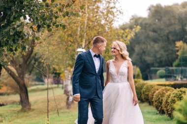 Gelinlik giymiş gelin ve takım elbiseli damat parkta el ele yürüyüp gülümsüyor.