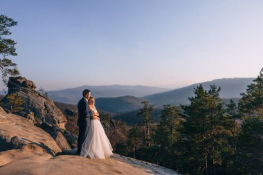 Gelin ve damat gün batımında kayaların arkasında dururlar. Yeni evliler gün batımında bir dağ ormanında