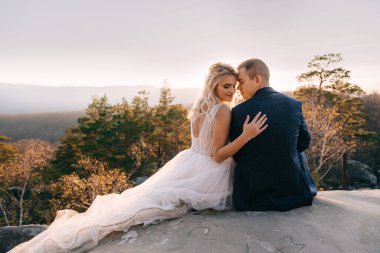 Genç bir çift yeni evli, gelin ve damat gün batımının, güzel dağların ve mavi gökyüzünün zemininde bir kayanın üzerinde oturuyorlar..