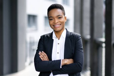 Güzel, genç, gülümseyen profesyonel siyahi kadın ofis binasının yanında duruyor. Elleri bağlı ve gülümsüyor.