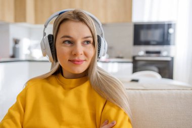 Gülümseyen, kulaklık takan, sarı bluzlu genç bir kadın şık bir ev mutfağında müziğin tadını çıkarıyor. Doğal ışık ortamı, modern rahatlık ve rahatlama anlarını vurgular, teknolojiyi konforla harmanlar..