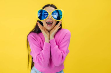 Heyecanlı pembe ve güneş gözlüğü takmış güzel kız sarı arka planda izole edilmiş indirimlere bakıyor.