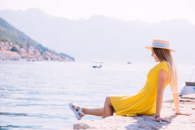 Sarı elbiseli neşeli bir kadın, deniz kenarında güneşlenerek oturur. Deniz kenarında güzel bir yaz günü, dinlenme ve doğanın güzelliğini kucaklar..