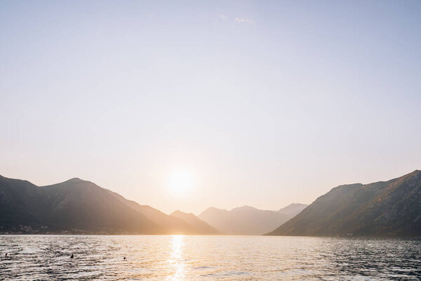 Яркое заходящее солнце над спокойным морем в окружении суровых гор, символизирующих мир и летнее спокойствие.