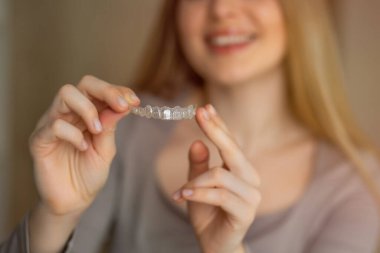 Bir kadın, elinde şeffaf bir ortodontik hizalama aleti tutarken arka planda gülümsüyor. Bu görüntü diş sağlığı, ortodonti ve modern diş hizalama çözümleri ile kendine güvenen bir gülümsemeyi temsil eder..