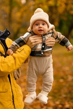 Sıcak bir kıyafet giymiş neşeli bir bebek bir sonbahar günü dışarıda bekletiliyor. Canlı sonbahar yeşillikleri büyüleyici bir mevsimlik arka plan sağlar, neşe ve bağlılık dolu anı vurgular.