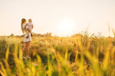 Çocuğunu taşıyan bir anne, günbatımının ve yaz atmosferinin tadını çıkarıyor. Uzun otlar ve kır çiçekleriyle çevrili..