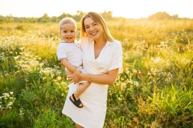 Neşeli bir anne gülümseyen oğlunu güneşli, yemyeşil bir çayırda tutuyor..