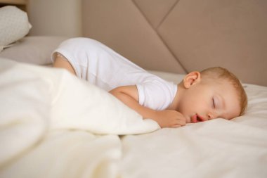 Genç bir çocuk, sakin bir atmosferdeki yumuşak battaniyelerle kaplı, rahat bir yatakta huzur içinde dinleniyor. Sahne sükunet, güvenlik ve çocukluğun masumiyetini sessiz bir uyku anında aktarır..
