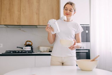 Parlak bir mutfakta neşeli bir kadın malzemeleri bir el çırpıcısıyla karıştırır ve pişirmeye hazırlanır..